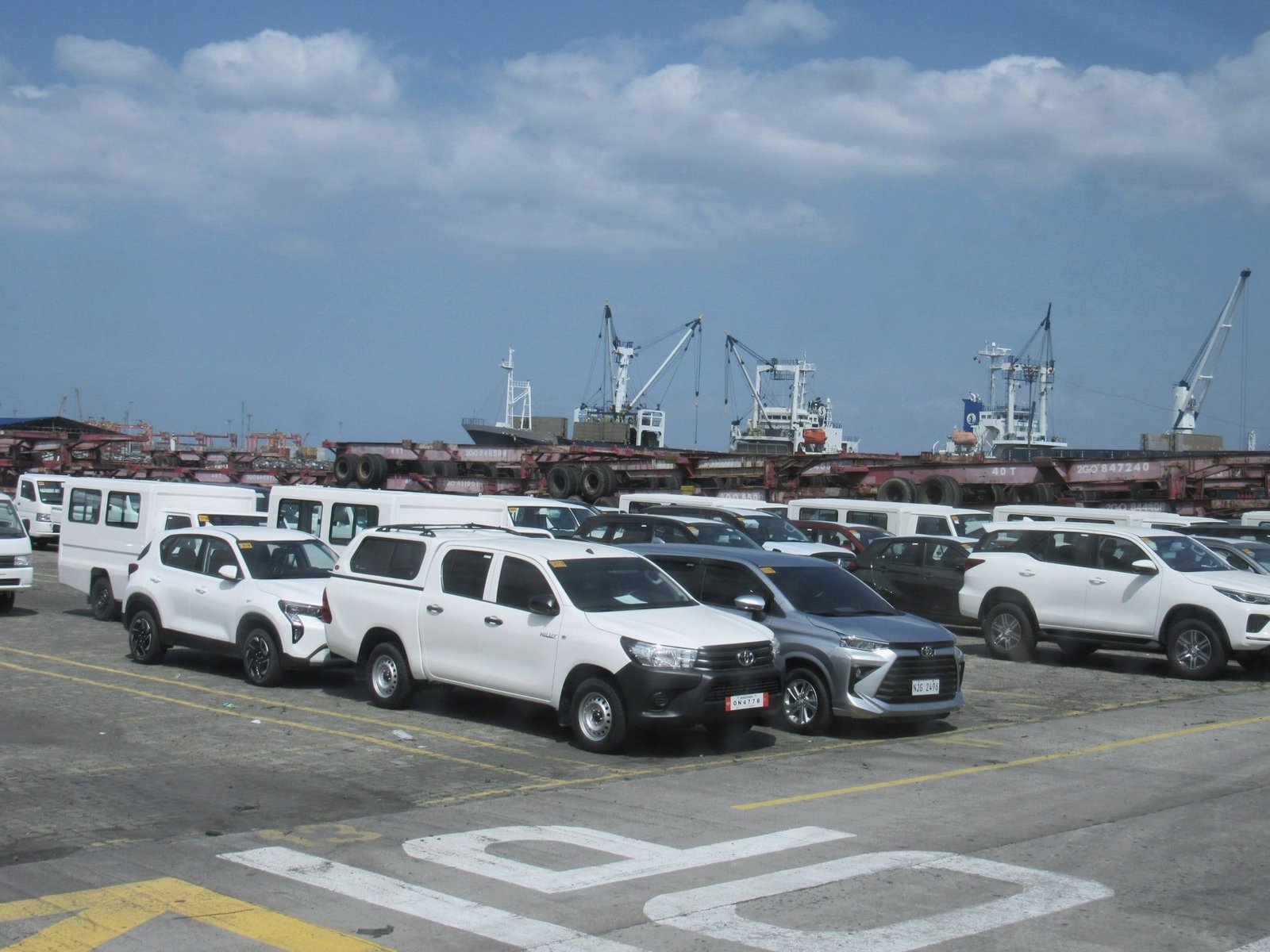 New vehicles lined up on a port apron with cargo ships behind